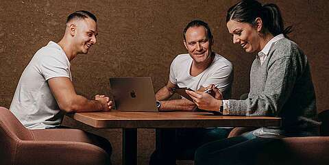 Menschen, die an einem Tisch sitzen. Eine Frau zeigt zwei Männern etwas auf einem Tablet.